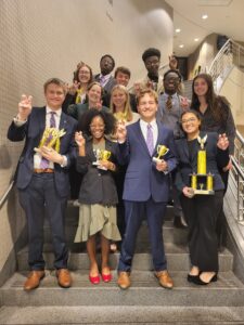 A group of college students smiling and posing with trophies in hand and celebratory hand gestures. Each of them is standing on a step on the stairs. Sharply dressed with formal business suits and dresses.
