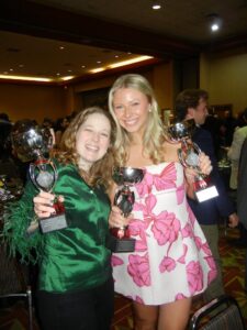 Ellie Griffin and Natalie Pelan stand side by side at an indoor awards event, smiling and holding up trophy cups in a banquet hall filled with other attendees.