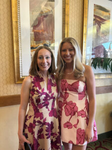 Two women stand side by side indoors, smiling at the camera, wearing pink floral dresses in front of framed artwork on a patterned wall.