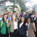 A group of well-dressed college students stand outdoors on campus, smiling and making celebratory hand gestures, dressed in business attire with trees and campus buildings in the background.