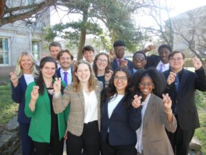 A group of well-dressed college students stand outdoors on campus, smiling and making celebratory hand gestures, dressed in business attire with trees and campus buildings in the background.