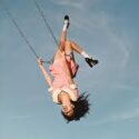 Olivia Rodrigo in a pink outfit swings upside down through the air on a rope against a clear blue sky, with her hair hanging downward and legs extended upward.