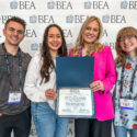 Four students pose in front of a wall with B-E-A repeated across it. Two of the students are holding a certificate in a folder.