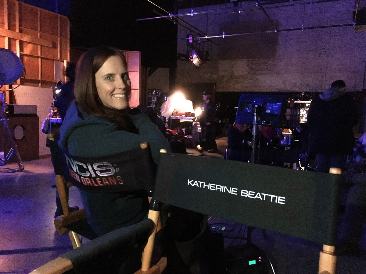 A woman sits in a directors chair with "NCIS: New Orleans" across the back. The directors chair next to her bears the name "Katherine Beattie."