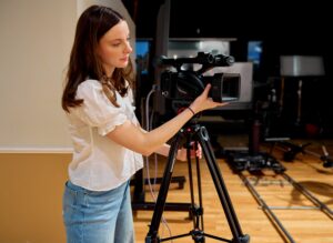 A person is operating a professional video camera on a tripod in a studio setting. The background includes lighting equipment and wooden floors.