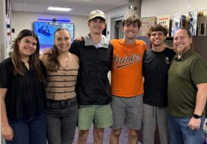 A group of six people is standing closely together, smiling in an indoor setting. One person is wearing an Orioles shirt. There is a screen displaying NASA in the background and walls with posters and pictures.