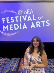 A young woman smiling and holding a glass award while standing in front of a large purple-lit screen that reads “BEA Festival of Media Arts.” She is wearing a floral dress and jewelry, and the setting appears to be an indoor event or awards ceremony with patterned carpet and stage lighting.