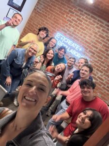 A group of people smiling and posing for a selfie in a room with brick walls. There is a neon sign in the background that reads "CREATE + COLLABORATE.