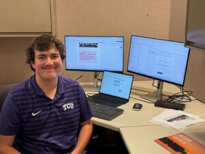 A person is sitting at a desk with two computer monitors and a laptop displaying various documents. They are wearing a purple TCU-branded shirt. A closed book titled "History Legacy" is on the desk. The setting appears to be an office or workspace.