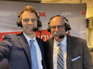 TCU Baseball alum Connor Wanhanen ’18 and Professor Chuck LaMendola pose together in a broadcast booth while wearing headsets and microphones during a live ESPN+ broadcast of a TCU baseball game. Wanhanen wears a navy suit with a light blue tie, while LaMendola wears a dark plaid jacket and striped tie. An ESPN+ and Big 12 backdrop is displayed behind them.