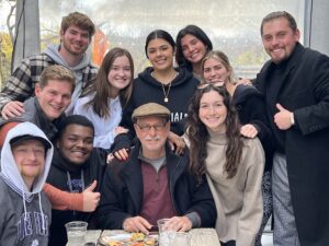 Professor Chuck LaMendola sits at the center of a group photo with students from his sports radio class gathered around him at an outdoor restaurant or patio setting. The group of smiling students poses closely together behind and beside him, with several giving thumbs-up gestures. LaMendola wears a flat cap, glasses, and a dark jacket over a maroon pullover while seated at a table with plates and drinks visible in the foreground.