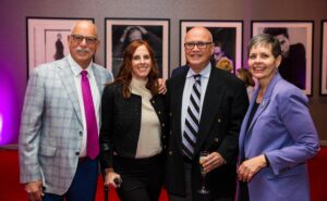Professor Chuck LaMendola, Katherine Beattie ’08, Professor Richard Allen, and Dean Kristie Bunton pose together at a formal indoor event with framed black-and-white portraits in the background. Chuck LaMendola wears a light plaid blazer with a bright pink tie, Katherine Beattie stands beside him in a black jacket and cream top, Richard Allen wears a dark suit and striped tie while holding a champagne flute, and Dean Kristie Bunton wears a lavender blazer and smiles toward the camera. Purple accent lighting and a red carpet add to the celebratory atmosphere.