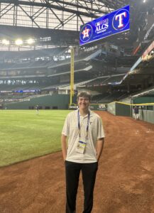 A person stands on the field inside a large baseball stadium. The roof structure is visible, and there is a scoreboard displaying the Texas Rangers and Houston Astros logos with "ALCS" written on it. The field is empty, and the lighting is bright.