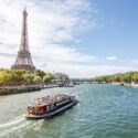 A daytime view of the Eiffel Tower in Paris, with a riverboat navigating the Seine River. The riverbanks are lined with green trees, and bridges are visible in the background under a clear blue sky.
