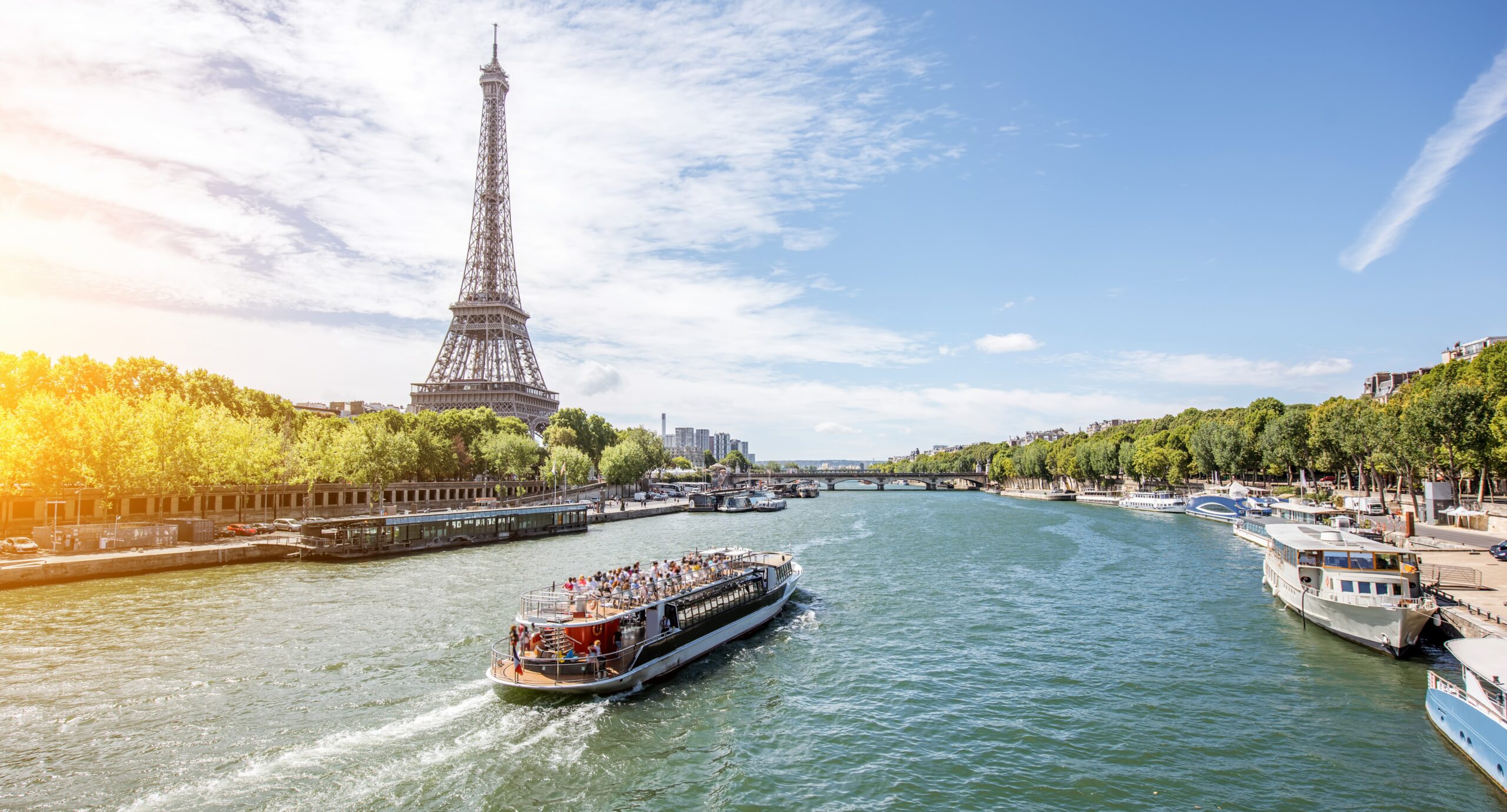 A daytime view of the Eiffel Tower in Paris, with a riverboat navigating the Seine River. The riverbanks are lined with green trees, and bridges are visible in the background under a clear blue sky.