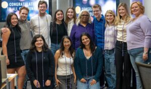 A group of people smiling and standing together indoors. Some are wearing honor cords. Two screens in the background display the message "Congratulations to All Our Winners."