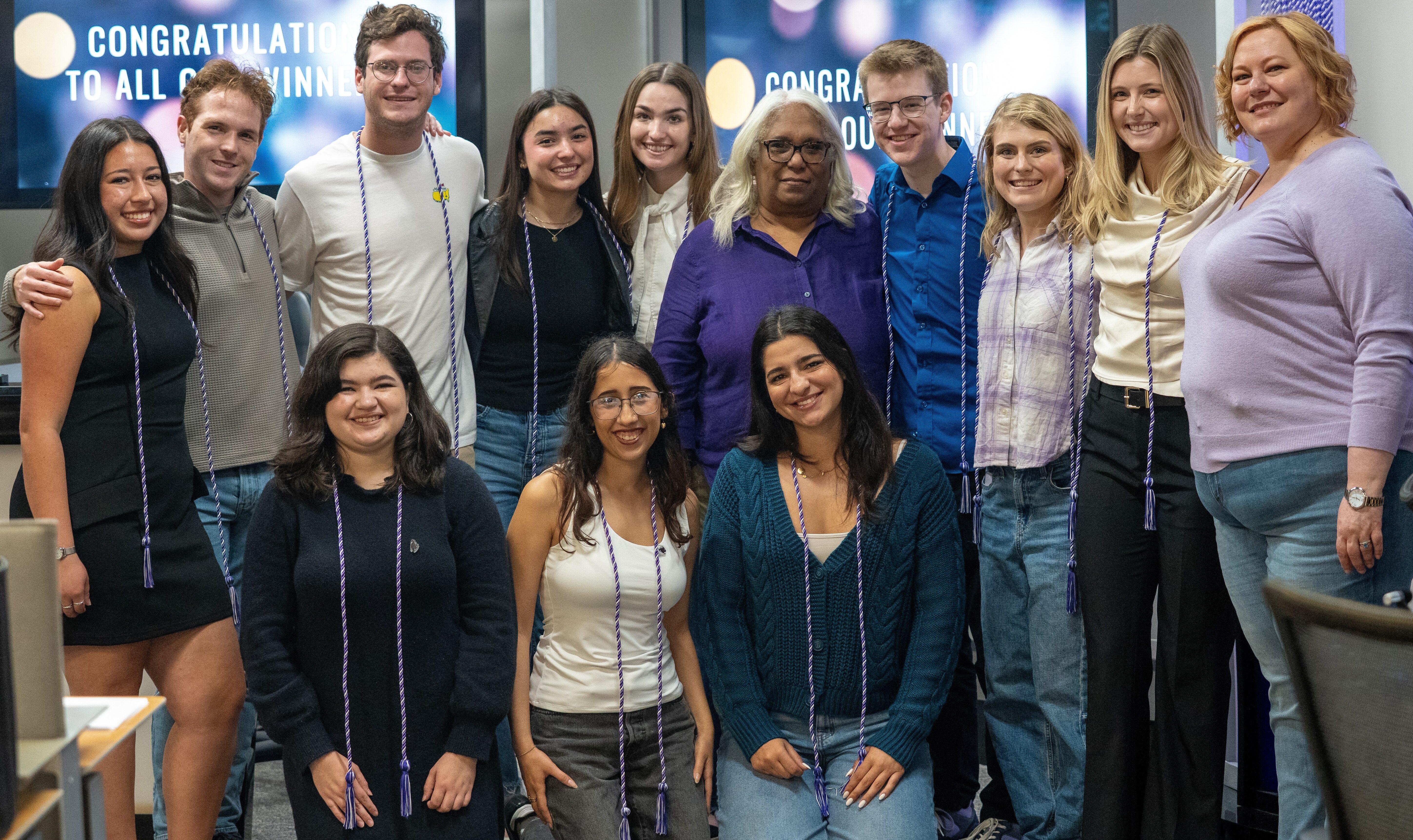 A group of people smiling and standing together indoors. Some are wearing honor cords. Two screens in the background display the message "Congratulations to All Our Winners."