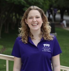 A person with curly hair is smiling outdoors. They are wearing a purple TCU Student Affairs polo shirt. The background includes greenery and a walkway.