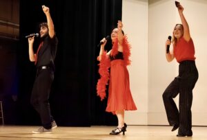 Three performers are on stage, energetically singing and holding microphones. They are dressed in red and black attire, with one in a red feather boa. The background features dark curtains and a white wall.