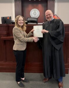 A person in judicial robes is shaking hands with another individual in a courtroom setting. They are both smiling and holding a certificate. Behind them is a wooden judge's bench with a seal.