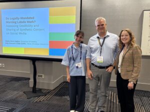 The image shows three individuals standing in front of a presentation screen. The screen displays a colorful slide with the title "Do Legally-Mandated Warning Labels Work? Assessing Credibility and Sharing of Synthetic Content on Social Media." The slide includes names and institutional affiliations: Alexis Shore, Syracuse University; Chip Stewart, Ellie Griffin, Texas Christian University.