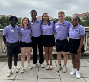 Five individuals pose together and smile. They are all wearing purple polos with a scenic background behind them.