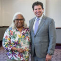 Two individuals stand together, one wearing a colorful outfit and the other in a suit.