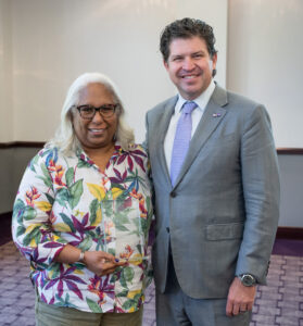 Two individuals stand together, one wearing a colorful outfit and the other in a suit.