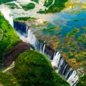 Aerial view of Victoria Falls, with water cascading down rugged cliffs into a gorge. Surrounding the falls are lush green landscapes and winding roads. The Zambezi River flows above the falls, dotted with small islands.