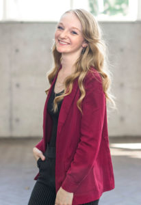 A young white woman with long curly blonde hair wearing a red jacket smiles at the camera.