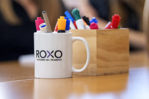 A coffee mug adorned with the Roxo logo and filled with pens and markers sits on a desk in the Roxo agency room.