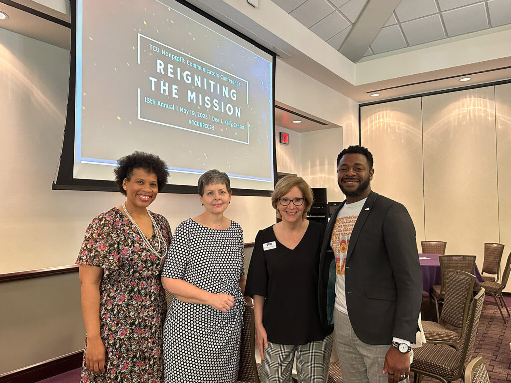Four people pose together in front of a digital screen in a conference room. The text on the screen reads " T C U nonprofit communicators conference. Reigniting the mission. 13th annual. May 19, 2023. Dee J Kelly Center. Hashtag T C U N P C C 23