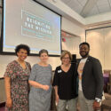 Four people pose together in front of a digital screen in a conference room. The text on the screen reads " T C U nonprofit communicators conference. Reigniting the mission. 13th annual. May 19, 2023. Dee J Kelly Center. Hashtag T C U N P C C 23