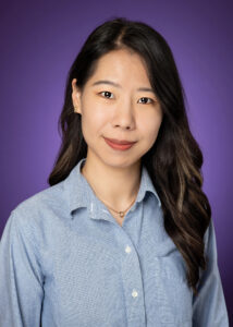 Professional headshot of a person with long dark hair wearing a light blue button-down shirt, posed against a purple background and looking at the camera with a slight smile. They are wearing small gold earrings and a delicate necklace.