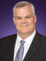 A gray-haired smiling white man in a charcoal suit, white shirt and purple tie.
