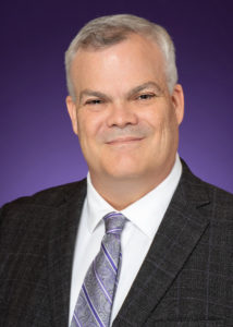 A gray-haired smiling white man in a charcoal suit, white shirt and purple tie.