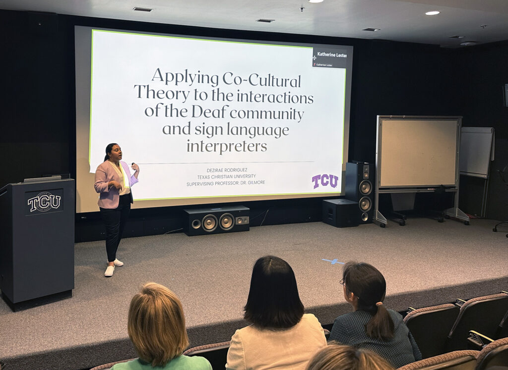 A student, standing on a stage, speaks to an audience. Behind her is a large screen. Text: Applying Co-Cultural Theory to the interactions of the Deaf community and sign language interpreters. 