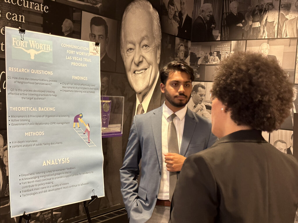 A student stands next to a large presentation poster and listens to another person facing away from the camera. 