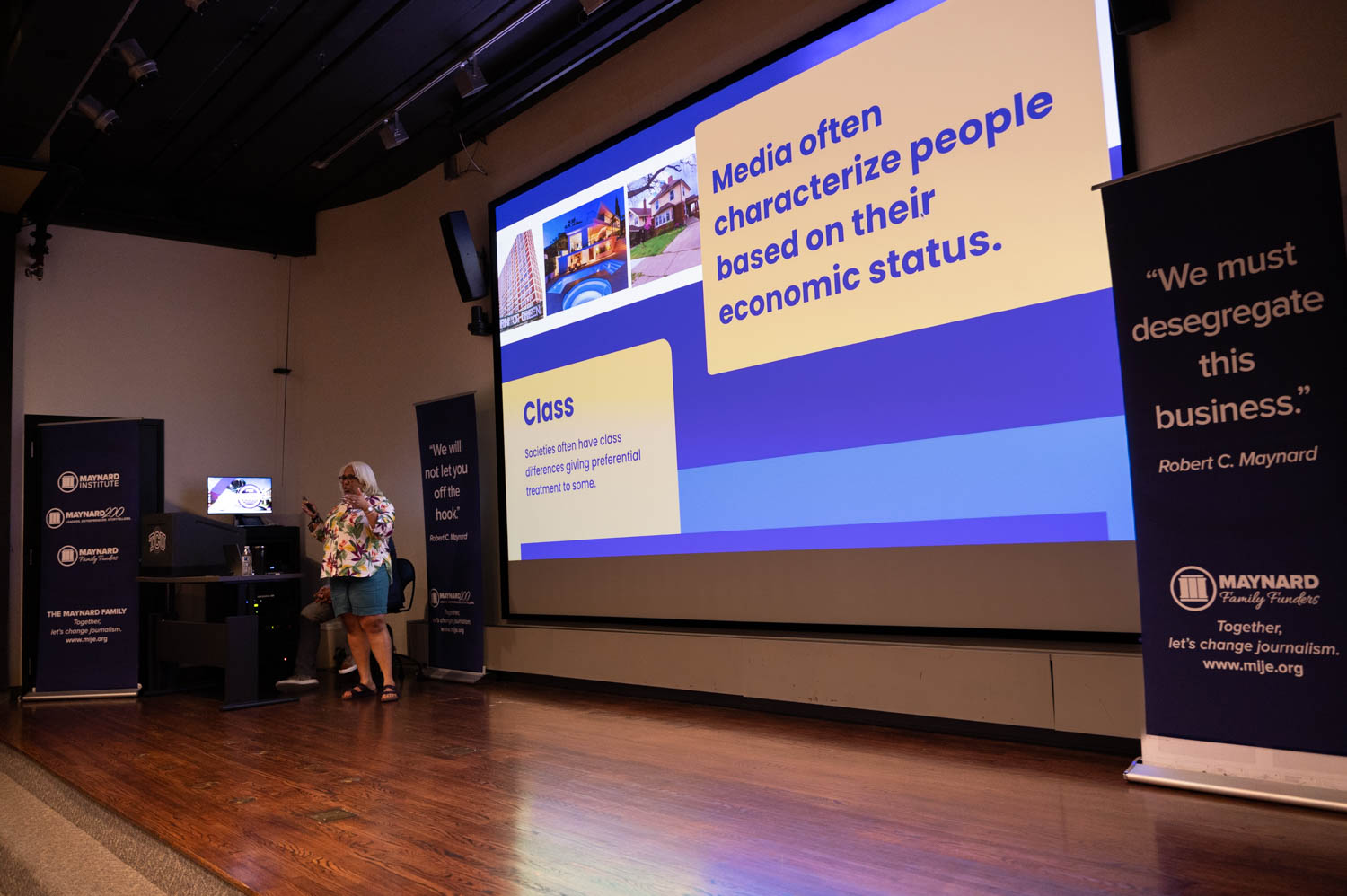 A professor speaks on stage in front of a screen with the text "Media often characterize people based on their economic status."
