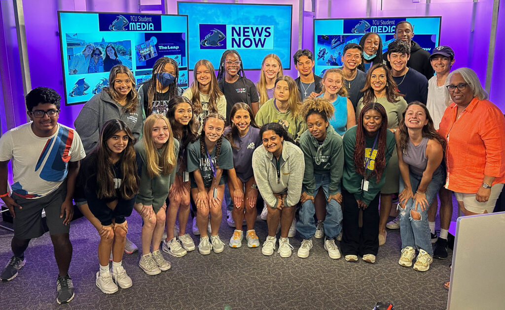 A group of high school students pose with their teacher and a journalist in a newsroom.