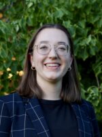 A person in a dark blue plaid blazer with a black shirt underneath is smiling. They are wearing glasses and earrings and are posed in front of a tree with deep green leaves and flecks of yellow throughout it.