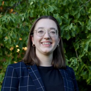 A person in a dark blue plaid blazer with a black shirt underneath is smiling. They are wearing glasses and earrings and are posed in front of a tree with deep green leaves and flecks of yellow throughout it.