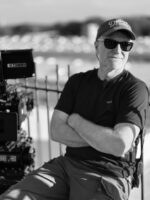 The image shows a person sitting beside a large film camera, outdoors, wearing sunglasses, a cap with the word "Dreams," and a headset. The background is blurred, featuring a railing and distant buildings.