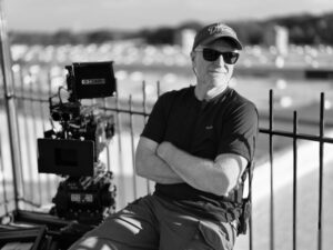 The image shows a person sitting beside a large film camera, outdoors, wearing sunglasses, a cap with the word "Dreams," and a headset. The background is blurred, featuring a railing and distant buildings.
