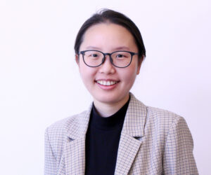 A person with glasses, wearing a checkered blazer and dark shirt, is smiling in front of a plain white background.