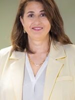 A person standing indoors, wearing a light-colored blazer over a blouse, with medium-length hair, in front of a blurred background.