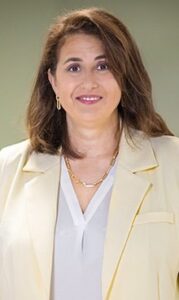 A person standing indoors, wearing a light-colored blazer over a blouse, with medium-length hair, in front of a blurred background.