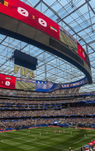A crowded soccer stadium in Los Angeles is shown with a large "FIFA" sign overhead. The field hosts a match between teams, and the scoreboard displays match details. The stadium is filled with spectators, and team flags are visible on the screens.