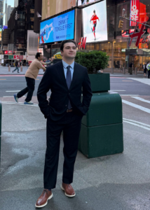 A person in formal attire stands on a city street with large digital billboards and an H&M store in the background. Tall buildings and pedestrians are visible, and one billboard displays an advertisement with the text "DRINK THRIVE FIRST." The scene takes place in a bustling urban area.