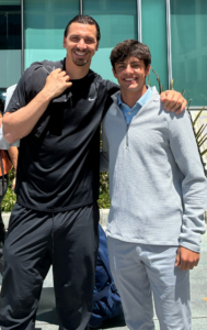 Two people are standing together outdoors in front of a building with glass windows. The person on the left is wearing black athletic attire, and the person on the right is wearing a light-colored long-sleeve shirt. Both are smiling and relaxed, with one person's arm around the other's shoulder.
